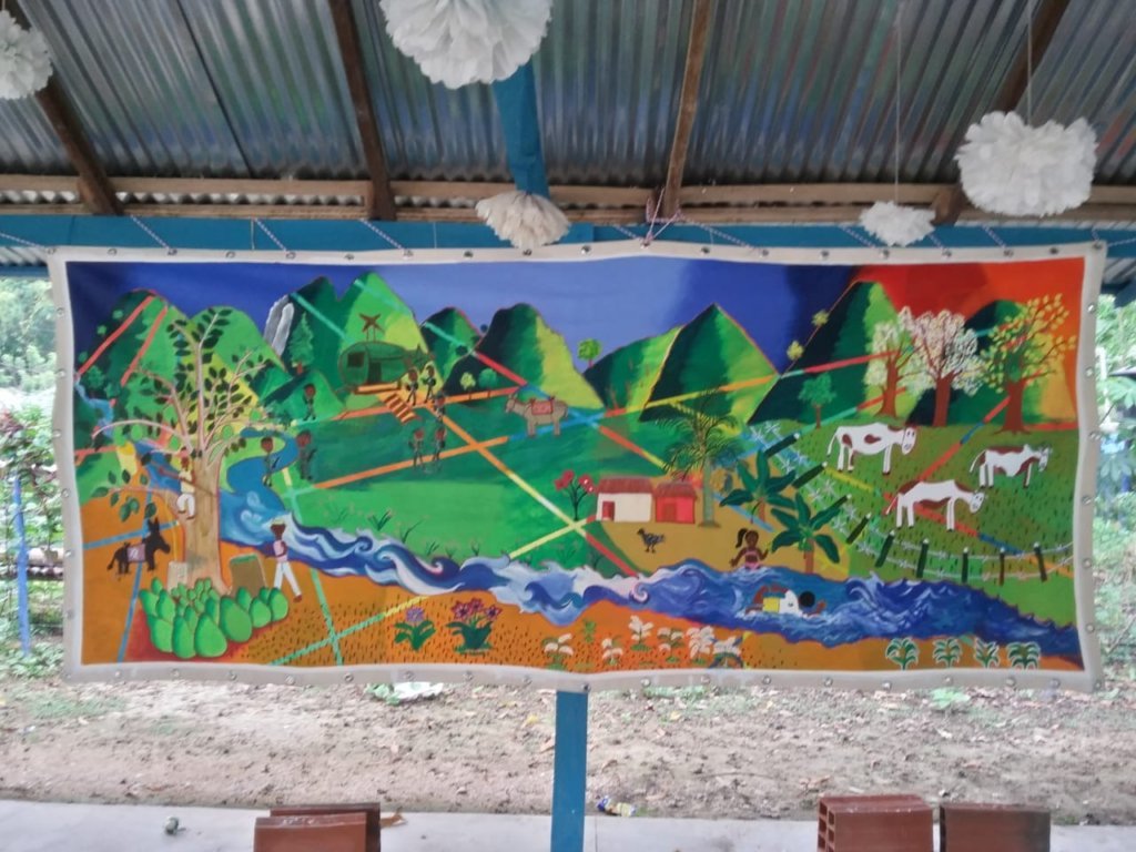 Mural en el corregimiento de Lázaro, en la zona de la Alta Montaña de El Carmen de Bolívar. Así lo describe un participante: “Donde está el palo de aguacate: ese era el pasado, cuando estaba la guerra, allí comienza el arroyo. El presente es donde están las casas, y donde están las vacas es el futuro que queremos”.